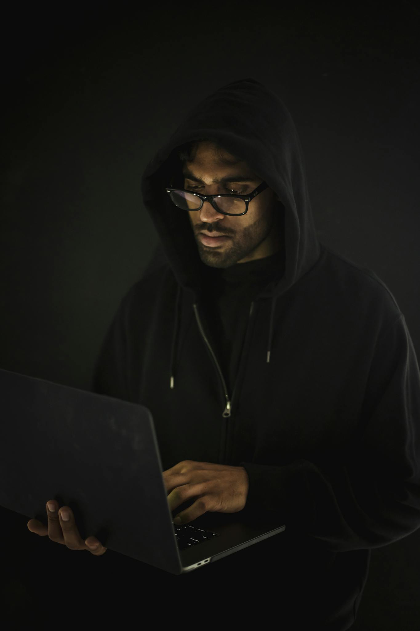 Serious young male in black hood and eyeglasses standing with netbook in hand and hacking database in dark room