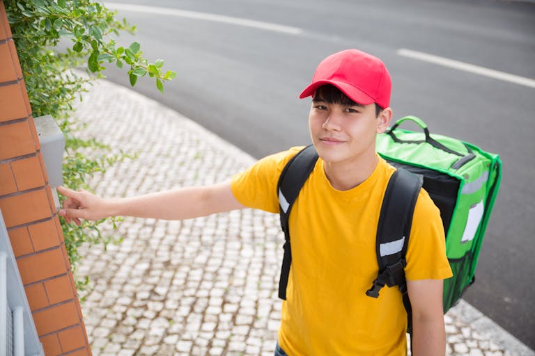 A cheerful kid in a red cap rings a doorbell, wearing a yellow shirt and carrying an insulated bag.